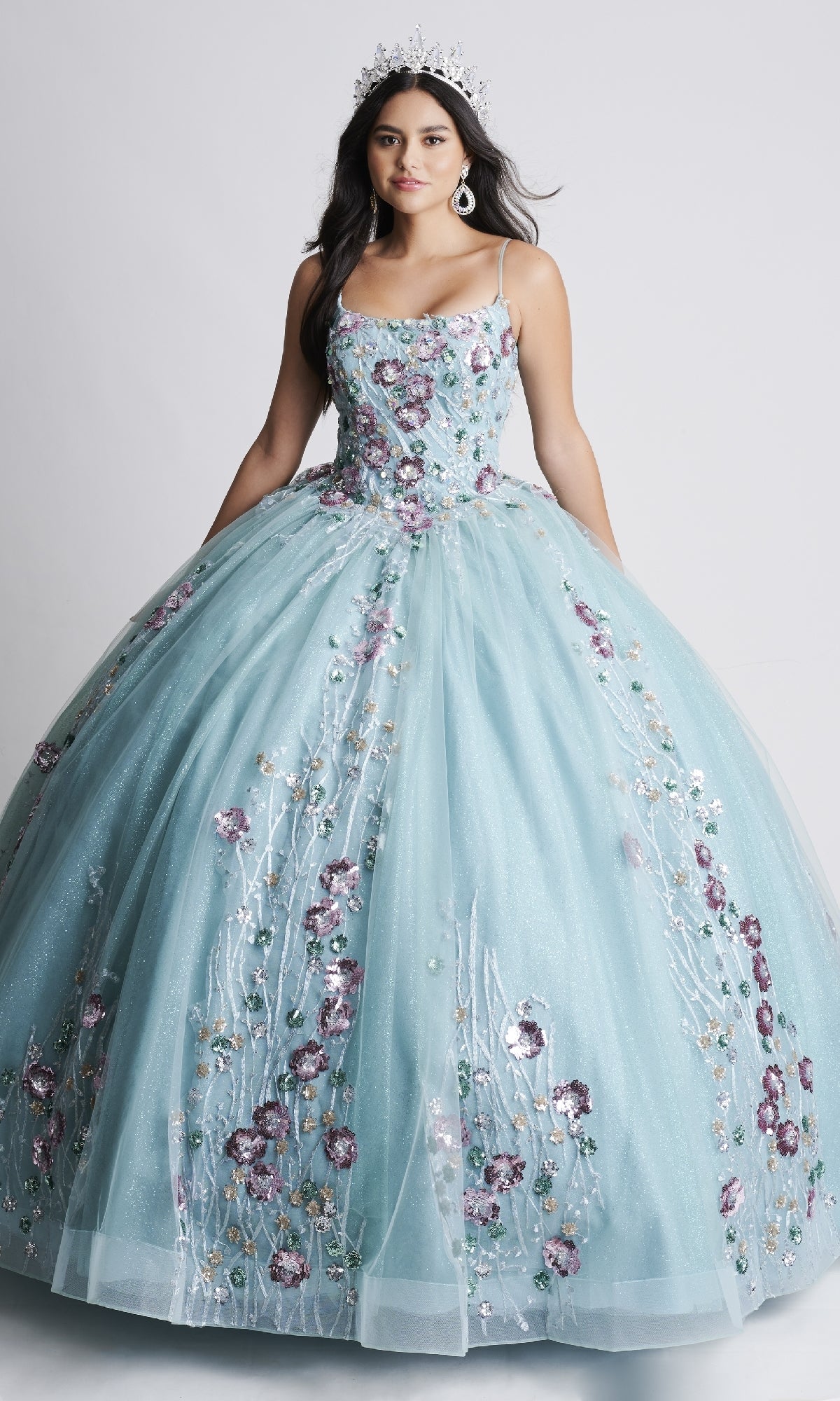 A young woman in the Nina Canacci Q105 long Quinceanera dress, a light blue ball gown with floral embroidery and a tiara, stands against a plain background.
